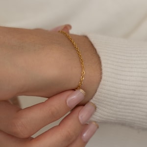 May include: A delicate gold chain bracelet. The bracelet is worn on a wrist, with the clasp being secured. The background is a white sweater. The bracelet is a simple, elegant piece of jewelry.