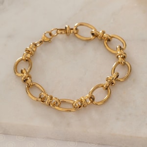 May include: A gold-colored bracelet with an oval link chain design. The bracelet is made of a shiny, polished metal and is displayed on a white marble surface. The bracelet is a piece of jewelry.