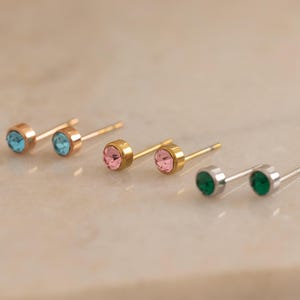May include: A collection of stud earrings with colored gemstones. The earrings are in gold and silver tones, with blue, pink, and green gemstones. The earrings are displayed on a light-colored surface.