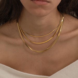 May include: A set of three gold necklaces, each with a different design, are shown. The necklaces are layered, including a herringbone chain, a flat chain, and a delicate chain. The jewellery is worn over a white top.