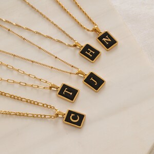 May include: Gold-toned necklaces with black rectangular pendants, each displaying a different letter in a white font. The chains have various designs, including rope and link styles. The pendants are centered on the chains.
