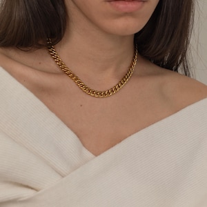 May include: A gold-colored chain necklace with a curb link design. The necklace is worn around the neck and rests on a white sweater. The chain has a shiny, metallic finish and is a classic jewelry piece.