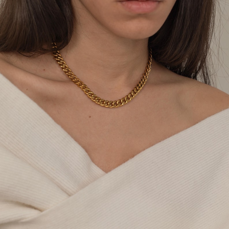 May include: A gold-colored chain necklace with a curb link design. The necklace is worn around the neck and rests on a white sweater. The chain has a shiny, metallic finish and is a classic jewelry piece.