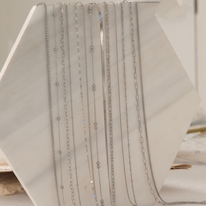 May include: A collection of silver necklaces displayed on a white marble surface. The necklaces feature various chain styles, including box, cable, and snake chains. The chains are delicate and shiny, reflecting the light. The background is a soft, neutral color.