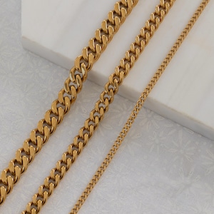May include: Gold-colored curb chain necklaces in varying widths. The chains are displayed on a white surface, showcasing their polished, reflective finish. The image highlights the different sizes and styles of the jewelry.