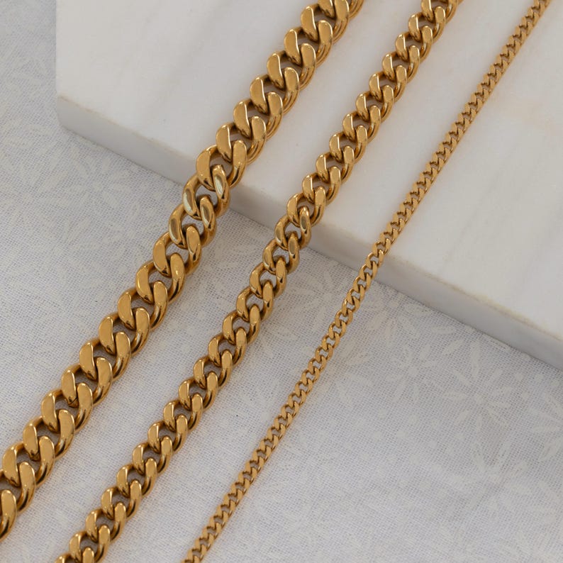 May include: Gold-colored curb chain necklaces in varying widths. The chains are displayed on a white surface, showcasing their polished, reflective finish. The image highlights the different sizes and styles of the jewelry.