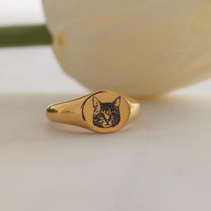 May include: A gold signet ring with a black cat face design. The ring has a polished finish and an oval face. The cat's face is etched into the gold, creating a detailed contrast. The ring is a classic piece of jewellery.