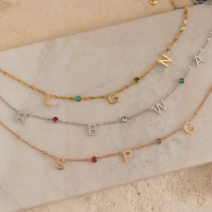 May include: Three gold, silver, and rose gold necklaces with initial charms and colorful gemstones. The necklaces are layered on a white marble surface.
