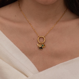 May include: A gold chain necklace with a charm ring and multiple charms, including a black heart, a green square, and a gold bee charm.