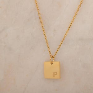 May include: Gold-colored necklace with a square pendant featuring the letter "P". The pendant hangs from a delicate chain. The background is a light, textured surface.