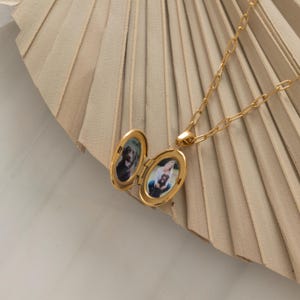 May include: Gold-colored oval locket necklace with a chain. The locket is open, revealing two small photos. The background is a beige, fan-shaped object. The necklace is a piece of jewelry.