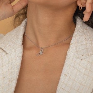 Pode incluir: Um colar de corrente de prata com um pingente de letra M em itálico.