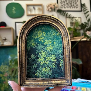 Puede incluir: Una obra de arte enmarcada con un diseño botánico en tonos verdes y amarillos. La pintura está dentro de un marco dorado en forma de arco. La obra de arte se sostiene en una mano, con una paleta de pinturas visible en primer plano.