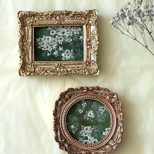 May include: Two ornate gold-toned frames, one rectangular and one circular, each containing a dark green print featuring small white flowers. The rectangular frame has intricate scrollwork, while the circular frame features a beaded inner border.
