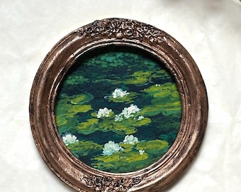 May include: A circular painting of white water lilies floating on a dark green pond, encased in an ornate, textured bronze-colored frame. The artwork features visible brushstrokes depicting lily pads and flowers in shades of green and white.