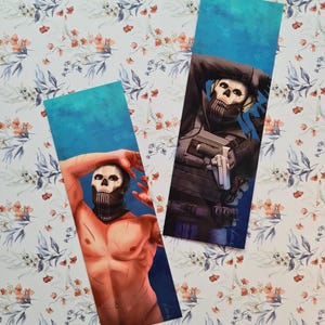 Peut inclure: Deux marque-pages rectangulaires avec des illustrations d'un personnage avec un masque de crâne. L'un montre le personnage torse nu, l'autre en équipement tactique. Les deux ont un fond bleu et sont posés sur un fond à motifs floraux.