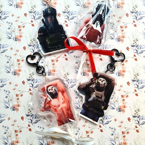 Puede incluir: Cuatro llaveros acrílicos transparentes con varios diseños de personajes. Cada llavero tiene un cierre en forma de corazón y un clip negro o dorado. Los llaveros están empaquetados en plástico transparente y atados con cintas rojas y blancas.