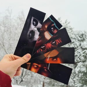May include: A hand displays five black rectangular stickers, each depicting a close-up of a face with detailed eyes and varying skin tones. The background is a snowy winter scene with bare trees.