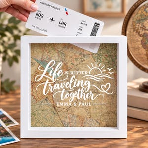 May include: A square white shadow box frame with a slot at the top, featuring a vintage map background and white text that reads Life is better traveling together and Emma & Paul, with a sun, wave, and heart graphic design printed on the glass.
