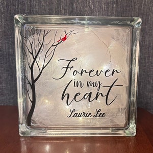 May include: A glass block with a white background and a black silhouette of a tree with a red cardinal perched on a branch. The text "Forever in my heart Laurie Lee" is written in black cursive lettering.