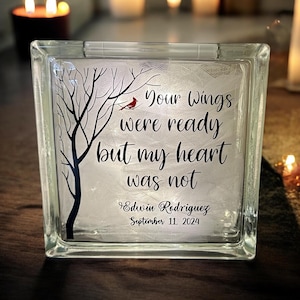 May include: A glass block with a white background and a black silhouette of a tree with a red cardinal perched on a branch. The text reads "Your wings were ready but my heart was not" with the name "Edwin Rodriguez" and the date "September 11, 2024" below.