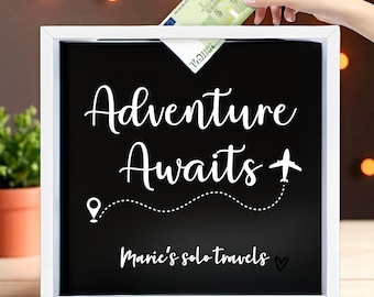 May include: A white-framed shadow box with a black interior, featuring the words "Adventure Awaits" in white script, a dotted line with a location pin, and a small airplane graphic. The text "Marie's solo travels" is also included. A hand is inserting a 100 Euro bill.
