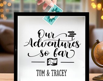 May include: A black shadow box with the words "Our Adventures so far" in elegant script. The names "TOM & TRACEY" are printed below. A hand is placing a postage stamp into the box, suggesting a travel or memory theme.