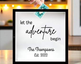 May include: A black shadow box with the words "let the adventure begin" and "The Thompsons Est. 2022" in black script. A hand is placing a teal and white postage stamp into the box.
