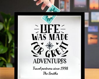 Peut inclure: Un coffret noir avec l'inscription "LIFE WAS MADE FOR GREAT ADVENTURES" en noir. Une main place un timbre-poste dans la boîte. Le texte "Travel partners since 1998 The Smiths" est en dessous. Un cadeau sur le thème du voyage.