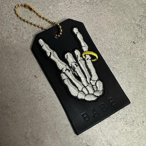 Puede incluir: Etiqueta de equipaje negra con una mano de esqueleto bordada en blanco haciendo un gesto de rock and roll. Un anillo dorado adorna el dedo anular. La etiqueta tiene una cadena dorada y la palabra "BABE" en relieve en la parte inferior.