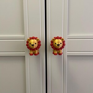 Pu&ograve; includere: Due pomelli per armadi a forma di leoni dei cartoni animati. I leoni sono dorati con criniere rosse e zampe rosse. I pomelli sono attaccati a un'anta bianca dell'armadio.