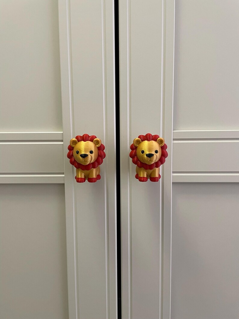 Pu&ograve; includere: Due pomelli per armadi a forma di leoni dei cartoni animati. I leoni sono dorati con criniere rosse e zampe rosse. I pomelli sono attaccati a un'anta bianca dell'armadio.