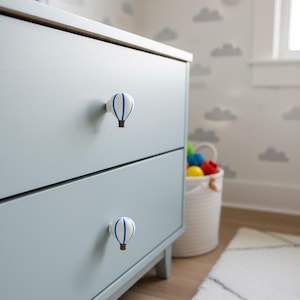 May include: A light blue dresser with two drawers, each adorned with a white and blue hot air balloon knob. The knobs have a brown base. The dresser is in a nursery setting with a cloud-patterned wallpaper and a white rug.
