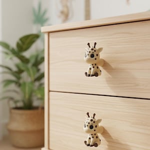 Op de afbeelding: Een lichtgekleurde houten dressoir met twee lades, elk versierd met een knop in de vorm van een giraf. De knoppen zijn crèmekleurig met zwarte vlekken en de giraf zit. Op de achtergrond staat een plant in een pot.