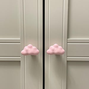 Op de afbeelding: Twee lichtroze wolkvormige kastknoppen op een lichtgrijze kastdeur. Deze decoratieve knoppen geven een speelse uitstraling aan elk meubelstuk. De knoppen zijn een charmante toevoeging.