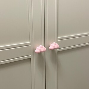 Op de afbeelding: Een close-up van een lichtgrijze kast met twee roze wolkvormige kastknoppen. De knoppen zijn gecentreerd op de kastdeuren en geven de neutrale meubels een speelse uitstraling.