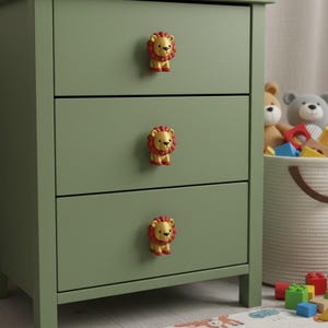 Pu&ograve; includere: Una cassettiera verde salvia a tre cassetti con pomelli a forma di leone. I pomelli a forma di leone sono gialli con criniere rosse. La cassettiera &egrave; in una cameretta con giocattoli sullo sfondo.