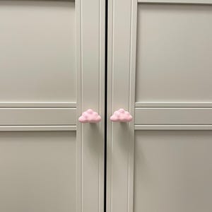 Op de afbeelding: Twee roze, wolk-vormige kastknoppen bevestigd op een lichtgrijze kast. De knoppen zijn gecentreerd op de deurpanelen, wat een speels element toevoegt. De kastdeuren zijn gesloten.
