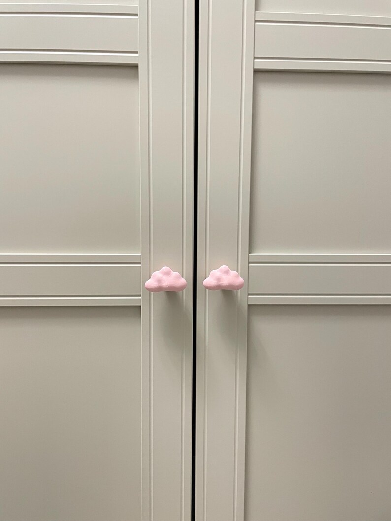 Op de afbeelding: Twee roze, wolk-vormige kastknoppen bevestigd op een lichtgrijze kast. De knoppen zijn gecentreerd op de deurpanelen, wat een speels element toevoegt. De kastdeuren zijn gesloten.