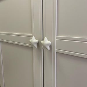 May include: A light grey cabinet with two doors, each featuring a white star-shaped handle. The doors have a recessed panel design with horizontal and vertical trim. The cabinet is a neutral colour.