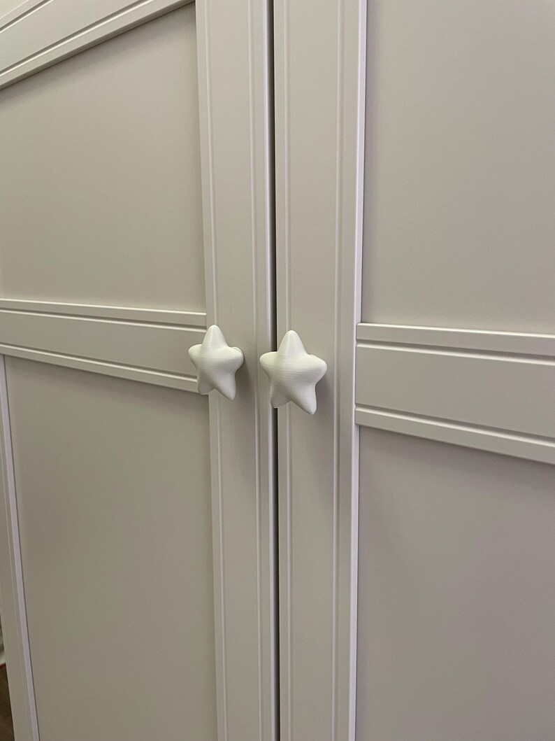 May include: A light grey cabinet with two doors, each featuring a white star-shaped handle. The doors have a recessed panel design with horizontal and vertical trim. The cabinet is a neutral colour.