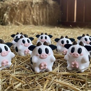 May include: A group of small, white cow figurines with black markings, ears, and horns, and pink udders and noses. The figurines are placed on a bed of hay, with more hay and a wooden structure in the background.
