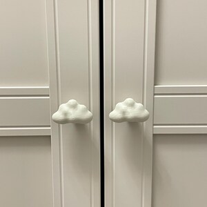 Może przedstawiać: Białe drzwi szafy z uchwytami w kształcie chmur. Drzwi szafy są pomalowane na biało i mają prosty, nowoczesny design. Uchwyty w kształcie chmur dodają szafie fantazyjnego charakteru.