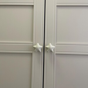 May include: A light grey cabinet with two white star-shaped door knobs. The cabinet doors have horizontal and vertical panel detailing. The star knobs are positioned centrally on the doors.