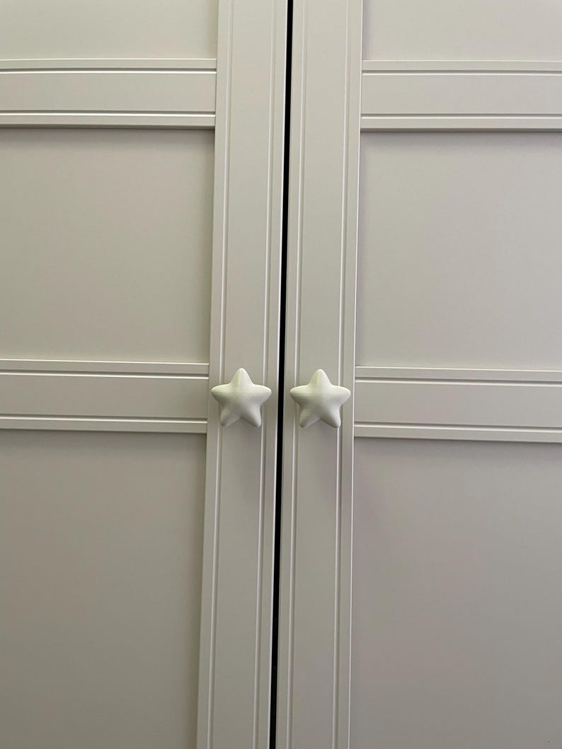 May include: A light grey cabinet with two white star-shaped door knobs. The cabinet doors have horizontal and vertical panel detailing. The star knobs are positioned centrally on the doors.