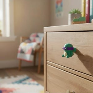 K&ouml;nnte beinhalten: Eine hellfarbene Holzkommode mit zwei Schubladen. Ein gr&uuml;ner und lila Cartoon-Charakter-Knauf ist an der oberen Schublade befestigt. Der Raum hat ein Fenster, einen Stuhl und einen bunten Teppich.