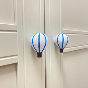 May include: Two white cabinet knobs shaped like hot air balloons with blue stripes and brown bases. The knobs are mounted on a light-colored cabinet door. The knobs are decorative and functional.