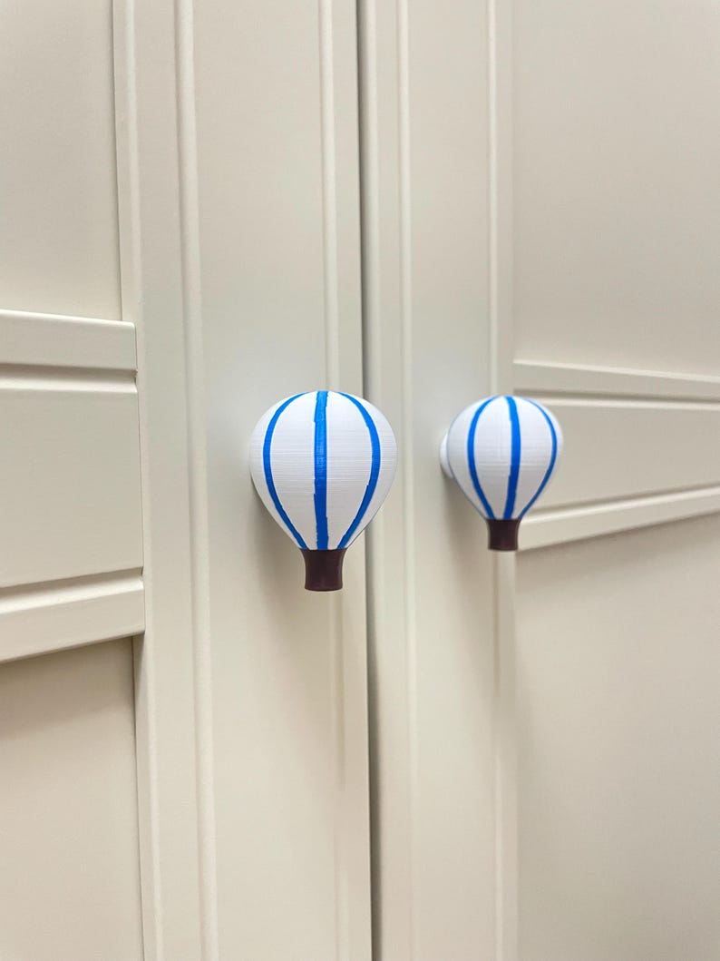 May include: Two white cabinet knobs shaped like hot air balloons with blue stripes and brown bases. The knobs are mounted on a light-colored cabinet door. The knobs are decorative and functional.