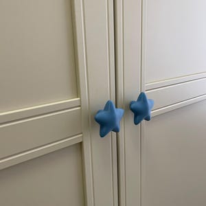 May include: A close-up of a white cabinet with two light blue star-shaped knobs. The knobs are centered on the cabinet doors, providing a decorative and functional element. The cabinet doors have a recessed panel design.