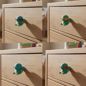 May include: Green turtle-shaped cabinet knobs with colored headbands, mounted on a light wood dresser. Each knob features a different colored headband: purple, orange, blue, and red. These decorative knobs add a playful touch to furniture.